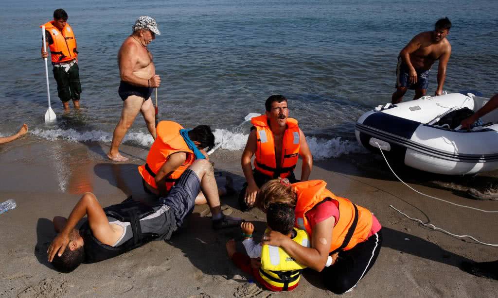 Ένας τουρίστας περνά δίπλα από έναν Ιρανό μετανάστη που καταρρέι δίπλα στο γιό του και τη γυναίκα του, λίγα λεπτά αφού έφτασαν στην ξηρά με μιά βάρκα χωρίς μηχανή.